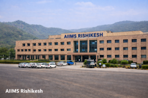 This image shows the main building of AIIMS Rishikesh (All India Institute of Medical Sciences) located in Uttarakhand, India.

The structure is a large, modern four-story building with a tan stone or tiled facade. The words "AIIMS RISHIKESH" are prominently displayed in blue lettering across the top center. The building is situated against a backdrop of the Himalayan foothills, characteristic of the Rishikesh region. In the foreground, there is a large paved area with several cars and a small bus parked near the entrance.