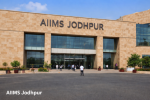 Front view of AIIMS Jodhpur hospital building with the “AIIMS JODHPUR” sign on a sandstone façade, glass entrance doors, people walking in and out, potted plants, and a clear blue sky.