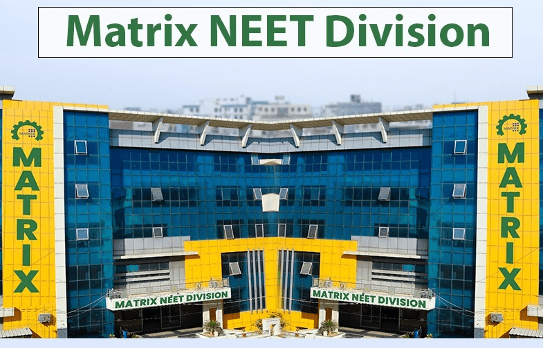 Front view of the Matrix NEET Division building. The modern building has blue-tinted glass windows with bright yellow accents. The words 'MATRIX' are vertically displayed on both sides of the building, and a banner above the entrance reads 'MATRIX NEET DIVISION'. The entrance is flanked by small plants, and the background shows a slightly blurred cityscape.