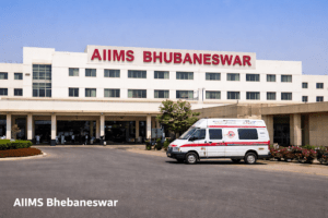 This image shows the main entrance of AIIMS Bhubaneswar (All India Institute of Medical Sciences) in Odisha, India.

The photo features a large, multi-story white medical building with the hospital's name displayed in prominent red letters at the top. In the foreground, there is an asphalt driveway with a white ambulance parked on the right side. The sky is clear and blue, and there is a small typo in the white caption text at the bottom left, which reads "AIIMS Bhebaneswar" instead of "Bhubaneswar."

AIIMS Bhubaneswar is a premier public medical research university and hospital, established in 2012 as part of the Pradhan Mantri Swasthya Suraksha Yojana (PMSSY).