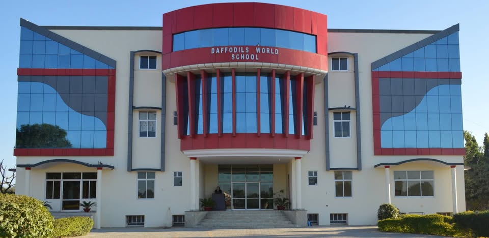 The Daffodils World School in Sikar boasts a striking and contemporary facade that immediately captures attention with its bold, modern architectural design under a clear blue sky. The central building features a prominent curved red canopy at the top emblazoned with "DAFFODILS WORLD SCHOOL" in large white letters, framed by expansive floor-to-ceiling blue-tinted glass windows that reflect the sky and create a dynamic, wave-like pattern across the structure. Symmetrical grey and beige panels accent the sides, while large angular red and grey geometric shapes add depth and visual interest, complemented by curved architectural elements at the base and roofline for a sleek, futuristic appeal. The grand entrance is accessed via wide steps leading to glass doors flanked by potted plants and small trees, with neatly trimmed green bushes lining the front, creating a welcoming and vibrant atmosphere. The overall campus exudes elegance and innovation, blending vibrant red accents with cool blue glass and neutral tones to symbolize creativity, growth, and a forward-thinking educational environment ideal for nurturing young minds in a bright, inspiring setting.