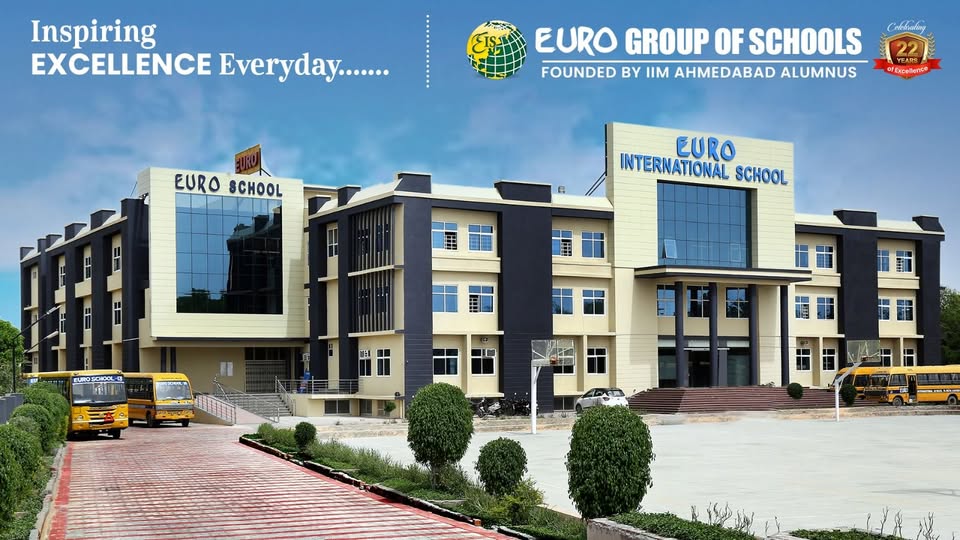 The Euro International School (part of the Euro Group of Schools, founded by IIM Ahmedabad alumni) in Sikar presents a striking, contemporary multi-building campus under a clear blue sky, featuring a sophisticated architectural design with a harmonious blend of beige-cream exterior walls, bold black vertical accents, large glass curtain windows for abundant natural light, and dark grey-black structural columns creating a modern, elegant facade. The prominent central block proudly displays "EURO INTERNATIONAL SCHOOL" in large blue lettering, flanked by adjacent sections labeled "EURO SCHOOL," with expansive glass panels reflecting the sky and surroundings for a premium, high-tech appearance. The foreground includes a wide, red-brick paved entrance pathway lined with neatly trimmed green shrubs and small trees, leading up to the main entrance with steps and railings, while several bright yellow school buses are parked prominently on both sides, ready for student transport, and a basketball hoop visible on the right adds to the emphasis on sports and extracurriculars. The overall scene radiates inspiration and excellence with lush greenery, open paved areas, parked vehicles including a car, and a well-maintained, spacious layout that highlights the school's 22 years of legacy, state-of-the-art facilities, and commitment to holistic education in a vibrant, welcoming environment.