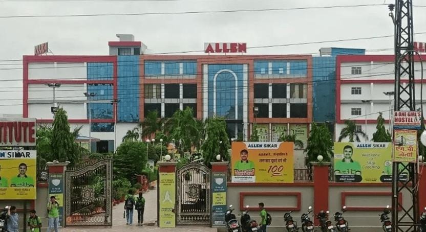 The image shows the impressive exterior of ALLEN Sikar coaching center, a multi-story modern building with a striking red, blue, and glass facade featuring a large prominent "ALLEN" sign at the top center. The entrance gate is visible with banners and hoardings displaying toppers' photos, motivational slogans like "लगभग 100 की" and student achievements, along with signs for "ALLEN Sikar" and "A One Girls Hostel." Palm trees line the pathway, students in uniforms walk toward the gate, and parked scooters/bikes are in the foreground under a cloudy sky, capturing the bustling yet disciplined vibe of this top NEET/JEE coaching institute in Sikar, Rajasthan.
