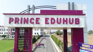 The image depicts the grand entrance archway of Prince Eduhub (also known as Prince Education Hub) in Sikar, Rajasthan, a prominent coaching and educational institute offering NEET, IIT-JEE, school, and competitive exam programs. The large overhead gate structure features a modern design with curved gray metal supports, a white top panel prominently displaying "PRINCE EDUHUB" in bold uppercase letters with "PRINCE" in red/pinkish-purple and "EDUHUB" in white, flanked by vertical purple pillars or accents on the sides. Below the arch, a wide paved driveway leads inward lined with green lawns and hedges, while a blue banner or sign in the foreground advertises "DEGREE + COMPETITION" or similar programs, capturing the welcoming yet disciplined gateway to this sprawling campus known for its lush green environment, multiple pathways from school to coaching excellence, and aspirational vibe under a clear sky.