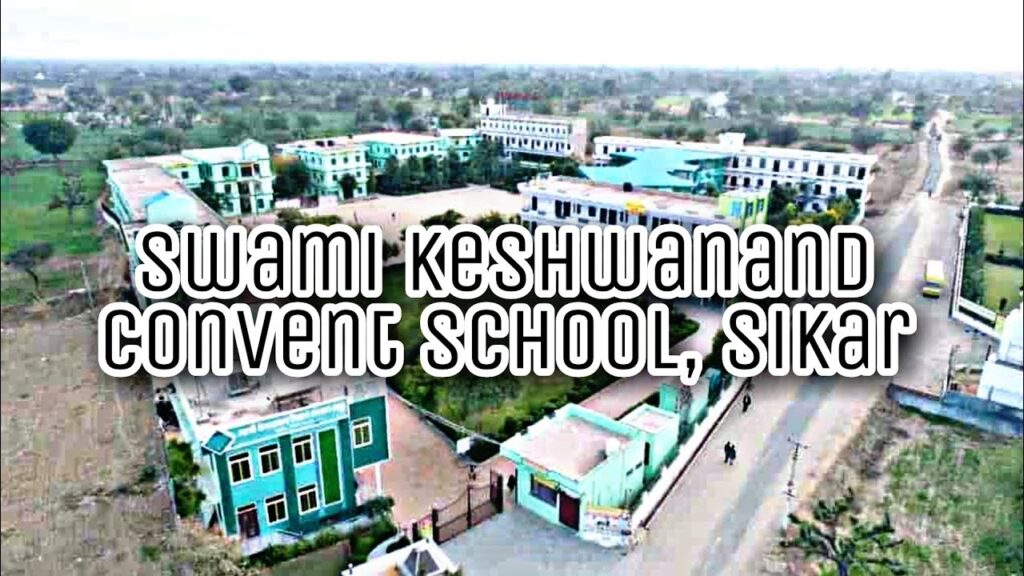 The Swami Keshwanand Convent School in Sikar offers a spacious and serene rural campus located at NH-52, Bhadhadar, surrounded by open arid fields, scattered trees, and village landscapes typical of Rajasthan's countryside, providing a peaceful, pollution-free environment away from urban hustle. The aerial view captures a well-organized complex of multiple multi-storey buildings painted in vibrant shades of turquoise blue-green (mint/aqua) with white accents, green-painted walls and roofs, greyish-white structures, and occasional colorful highlights like blue rooftops or signage, featuring rectangular blocks arranged around central open grounds, playgrounds with marked courts (such as basketball or multi-purpose areas), pathways, boundary walls with gated entrances, and scattered vehicles or people adding scale. The premises include clusters of academic blocks, hostels, administrative areas, and green patches with trees for shade, creating a welcoming, expansive setup that emphasizes holistic education in a natural, community-oriented setting with modern yet traditional architecture suited for boarding and day scholars from Pre-Primary to higher classes.