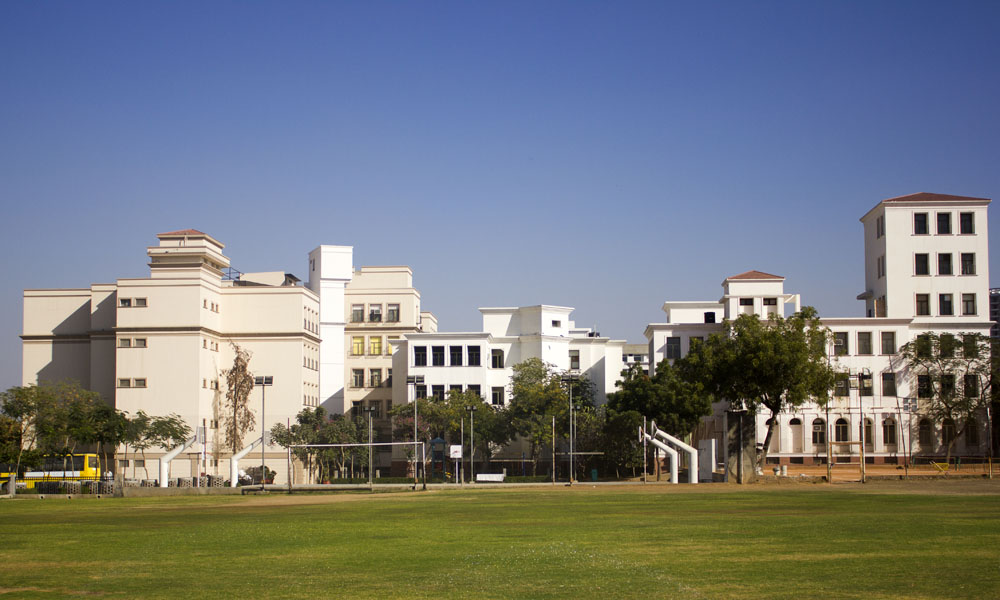 This photograph captures a wide panoramic view of Neerja Modi School in Jaipur, showcasing its impressive modern campus under a clear blue sky. The sprawling complex consists of multiple interconnected multi-storey buildings in elegant off-white and cream tones, featuring clean contemporary architecture with large rectangular windows, flat and gabled roofs in terracotta, subtle yellow accents, and white vertical elements. In the foreground lies a vast, well-maintained green sports field with lush grass, bordered by mature trees, tall floodlight poles, volleyball posts, and a yellow school bus parked near the buildings, creating a bright, spacious, and vibrant educational environment typical of a leading day and boarding school in Jaipur.