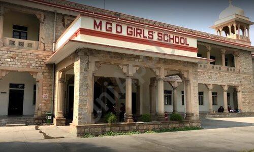 This image displays the grand main entrance of Maharani Gayatri Devi Girls’ School (MGD Girls’ School) in Jaipur, featuring a majestic colonial-era sandstone building with intricate stonework. The prominent portico is supported by thick carved pillars and rounded arches, leading into the interior, while a large white signboard with bold red lettering clearly reads “M G D GIRLS’ SCHOOL” across the top. The multi-storey structure includes balconies with railings, arched windows, and traditional Indian architectural details, with a small domed tower visible on the right side. In the foreground lies a spacious paved courtyard with a few small plants at the base of the entrance, and a couple of people can be seen sitting in the shaded background on the right, capturing the historic and elegant ambiance of the renowned all-girls boarding school.