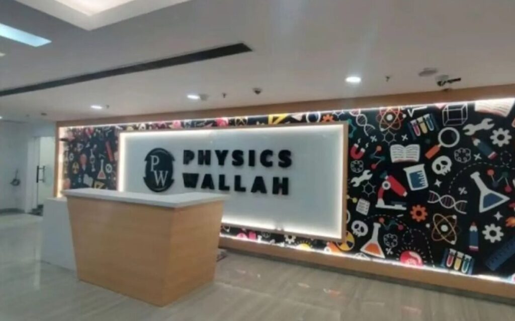 The photograph captures the bright and modern reception area of Physics Wallah (PW), also known as Physics Wallah Wallah, a popular edtech platform and coaching institute focused on JEE, NEET, and other competitive exam preparations. At the center stands a sleek, illuminated white reception desk with backlit signage prominently displaying "PHYSICS WALLAH" in bold black lettering, accompanied by the distinctive "PW" logo in a circular emblem to the left. Behind the desk, a large, eye-catching wall features glowing backlighting and is covered in a vibrant, colorful mural on a dark background, filled with playful scientific and educational icons such as test tubes, beakers, microscopes, DNA strands, atoms, books, gears, skulls, wrenches, light bulbs, periodic table elements, and various lab equipment scattered in an energetic, doodle-like pattern. The overall interior exudes a contemporary, motivational vibe with clean white ceilings, recessed lighting, and a spacious lobby atmosphere typical of PW's offline centers, emphasizing science, innovation, and student success.