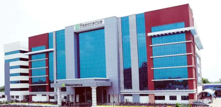 The image showcases the impressive corporate headquarters building of Resonance Eduventures in Kota, Rajasthan, a renowned coaching institute for JEE (Main & Advanced), NEET, and other competitive exams. The modern, multi-story structure features a striking facade with bold vertical color bands in red, blue, grey, and white, accented by large reflective blue-tinted glass panels that dominate the central and side sections, creating a sleek and contemporary architectural look. Prominent signage reading "Resonance" appears at the entrance canopy and atop the building in green lettering, with additional "Resonance" branding visible on the lower levels. The design includes symmetrical wings, overhanging elements, and a spacious foreground with greenery, parked vehicles, and a driveway, emphasizing its status as one of the prominent and well-maintained coaching campuses in Kota under a clear sky.
