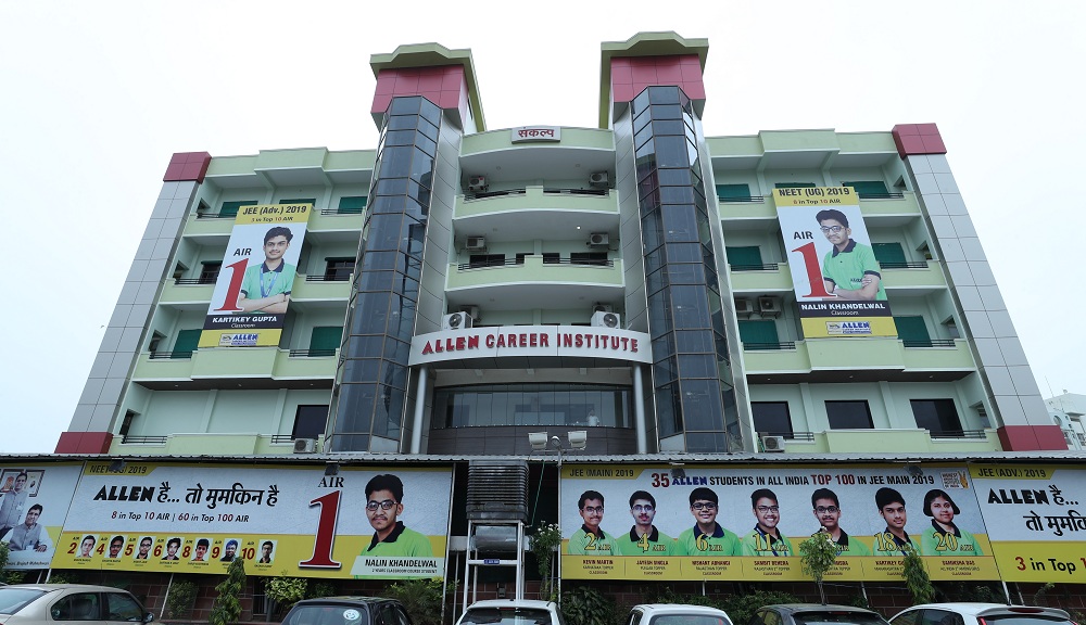 The photograph shows the multi-story building of ALLEN Career Institute in Jaipur, featuring a modern facade with glass panels, air conditioning units, and a prominent signboard reading "ALLEN CAREER INSTITUTE" at the entrance. The structure is adorned with large, colorful promotional banners and hoardings celebrating the institute's top performers from exams like JEE and NEET (particularly highlighting 2019 results), including massive posters of All India Rank (AIR) 1 achievers such as Nalin Khandelwal in NEET and others in JEE, along with lists of multiple students in the top 10, 20, and 100 ranks. These vibrant green-and-yellow banners cover the lower levels and surroundings, proclaiming slogans like "ALLEN है... तो मुक़म्मल है" and showcasing photos of smiling toppers in uniforms, creating a motivational and achievement-focused atmosphere typical of competitive coaching centers in India, with parked cars visible in the foreground under an overcast sky.
